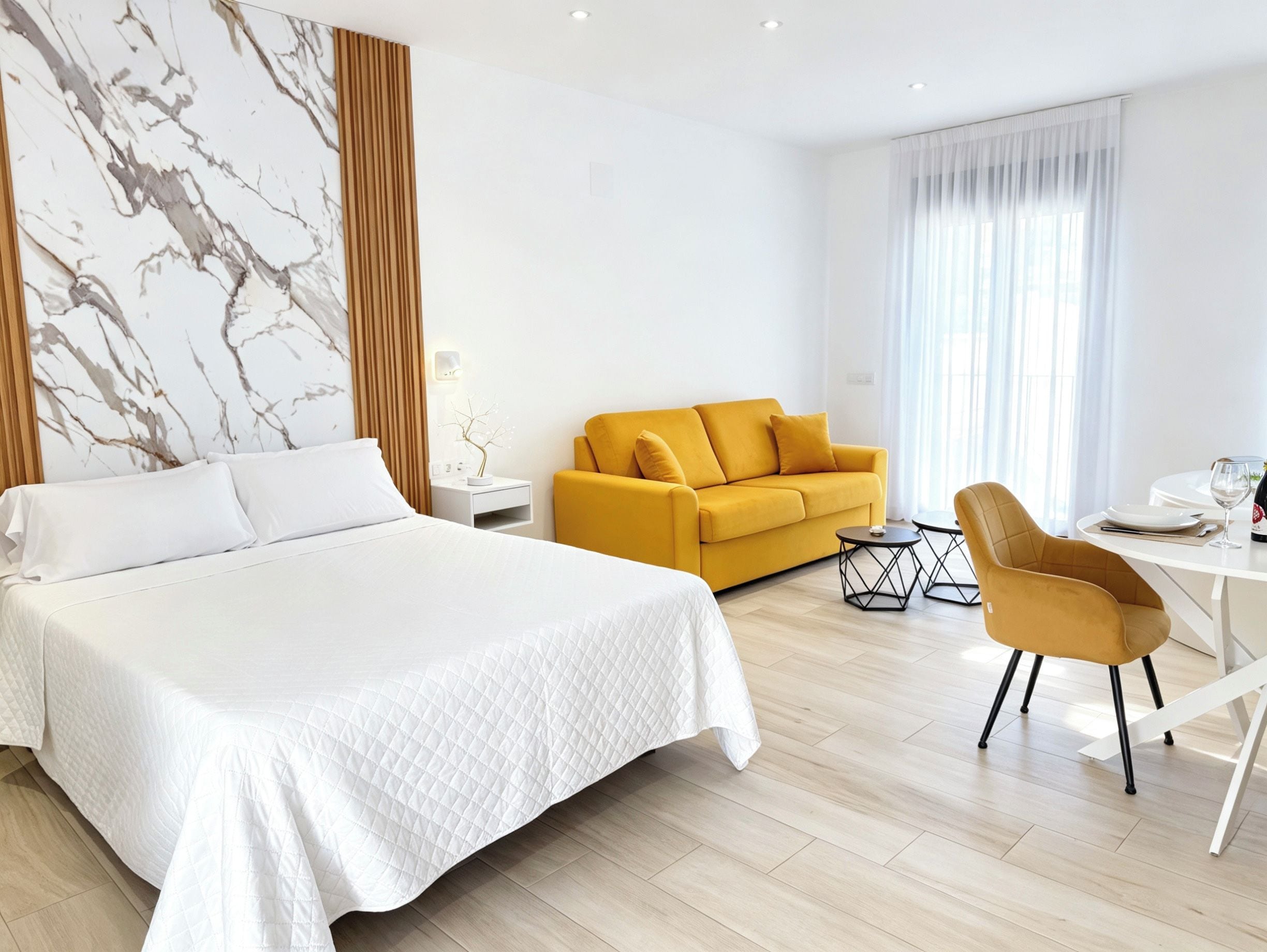 A hotel room with a large bed, a sofa, a dining table with chairs, and a window with curtains.