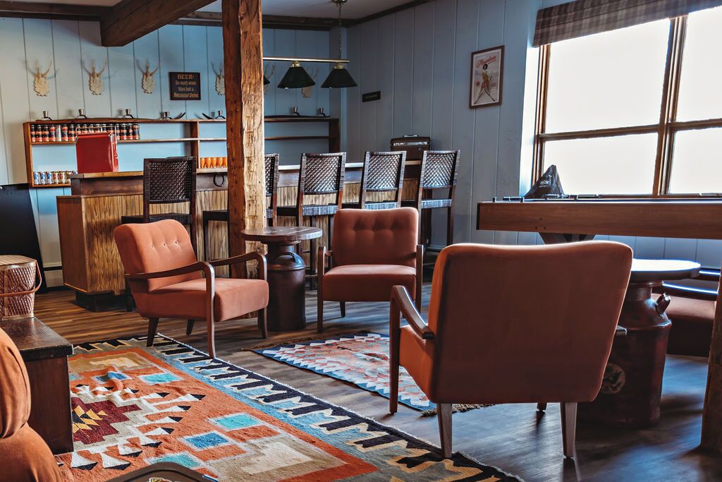 A cozy room with a wooden floor, a patterned rug, and orange chairs. There is a bar area with shelves and a sign, and a window with a view.