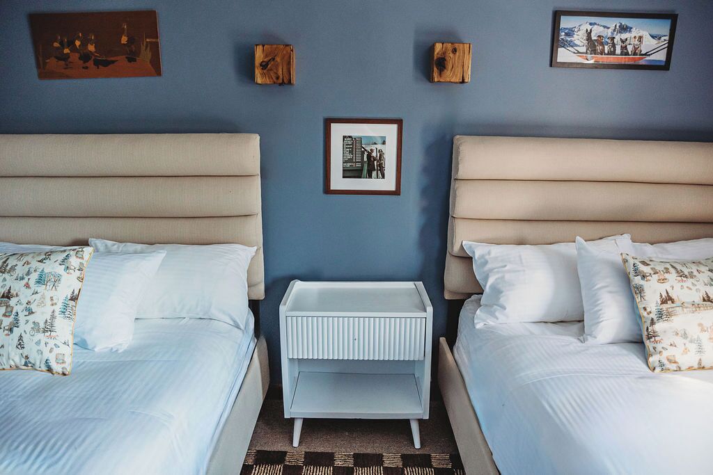 A hotel room with two beds, a small white nightstand, and a blue wall with framed pictures.