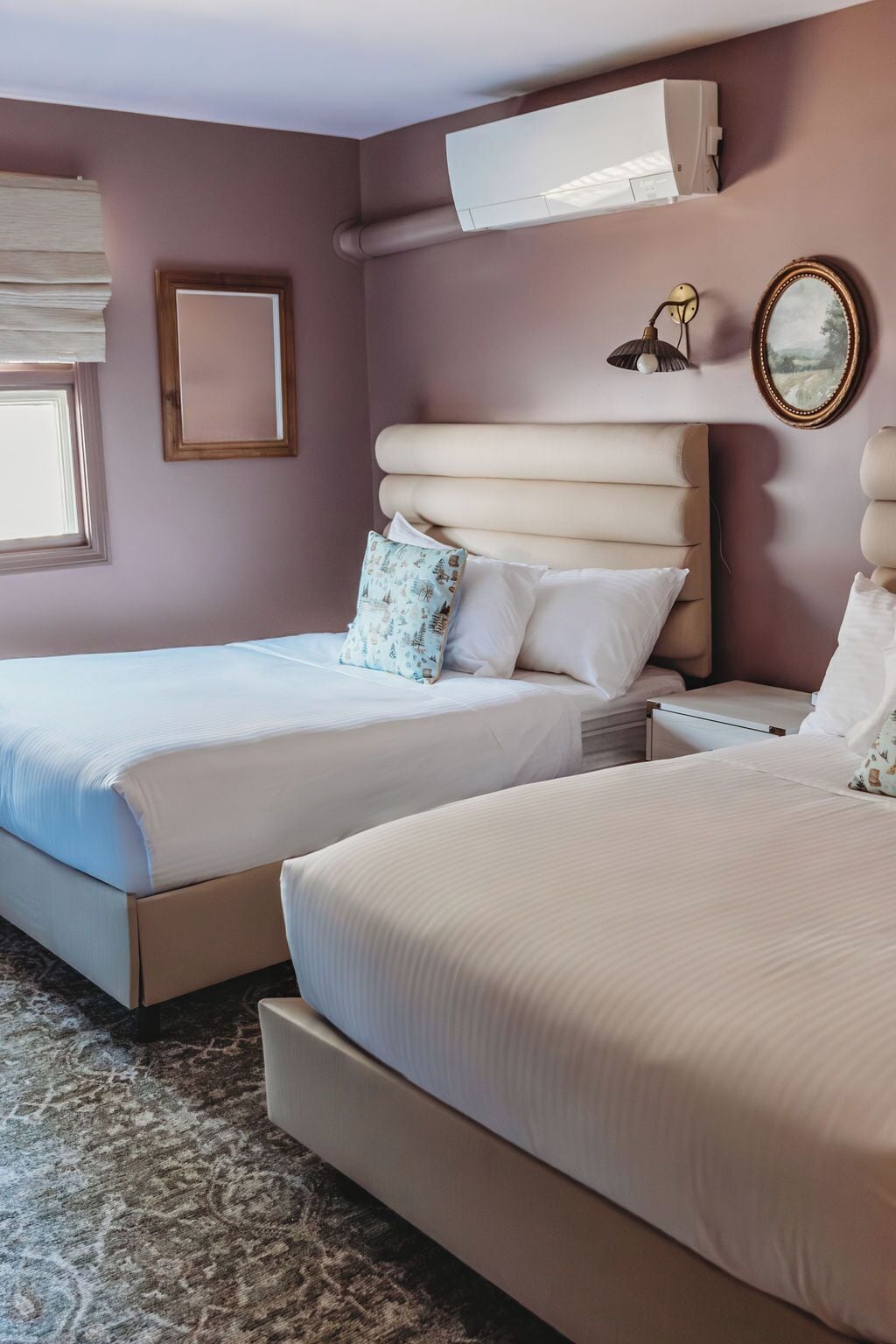 A hotel room with two beds, a large window with blinds, a wall-mounted air conditioning unit, and a bedside table with a lamp.