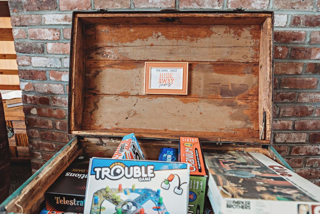 A wooden chest with a sign that reads "THE GAME CHESS HIDE AWAY" and various board games like "Trouble Game" and "Candyland" in front of it.