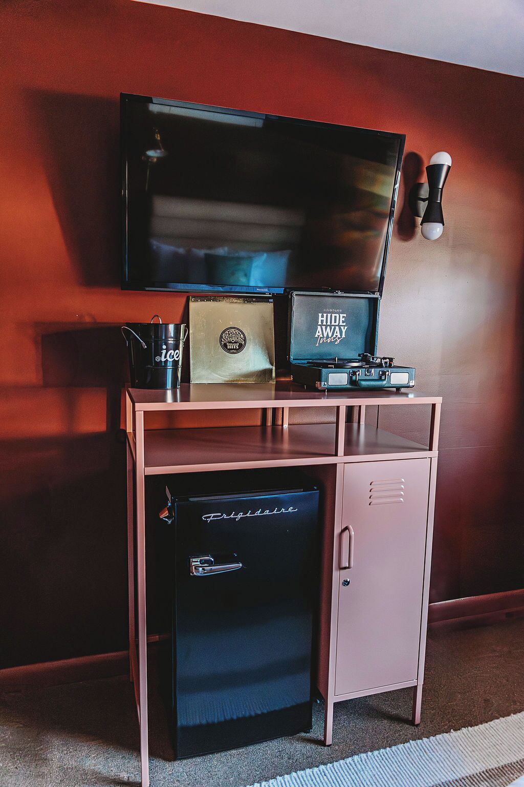 Una habitación compacta con un televisor montado en la pared, un refrigerador pequeño, una cafetera y un letrero que dice 'Hide Away'.