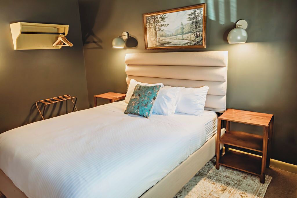 A neatly made bed with a headboard, two bedside tables, a wall-mounted air conditioner, and a painting above the bed.
