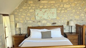 Un lit en bois dans une pièce aux murs de pierre, une table de chevet avec une lampe et une fenêtre avec des rideaux.