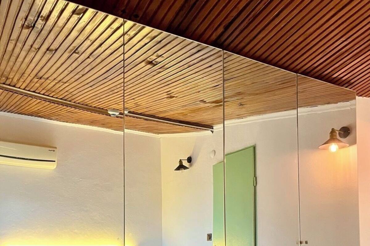 A modern hotel room with a wooden ceiling, a bed with white linens, a green door, and a wall-mounted air conditioner.