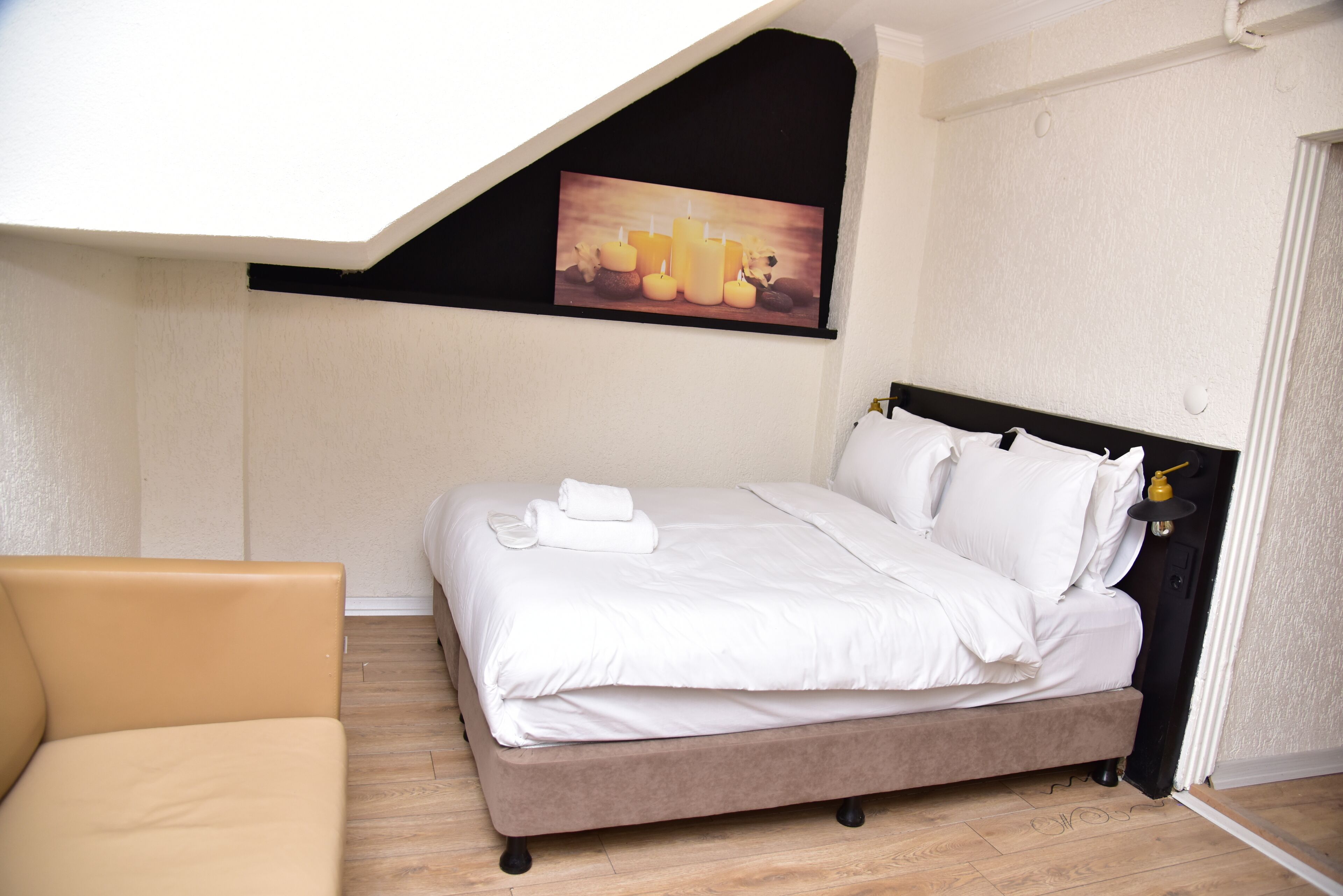 A neatly made bed with white linens and a folded towel, a tan armchair, and a painting of candles on the wall.