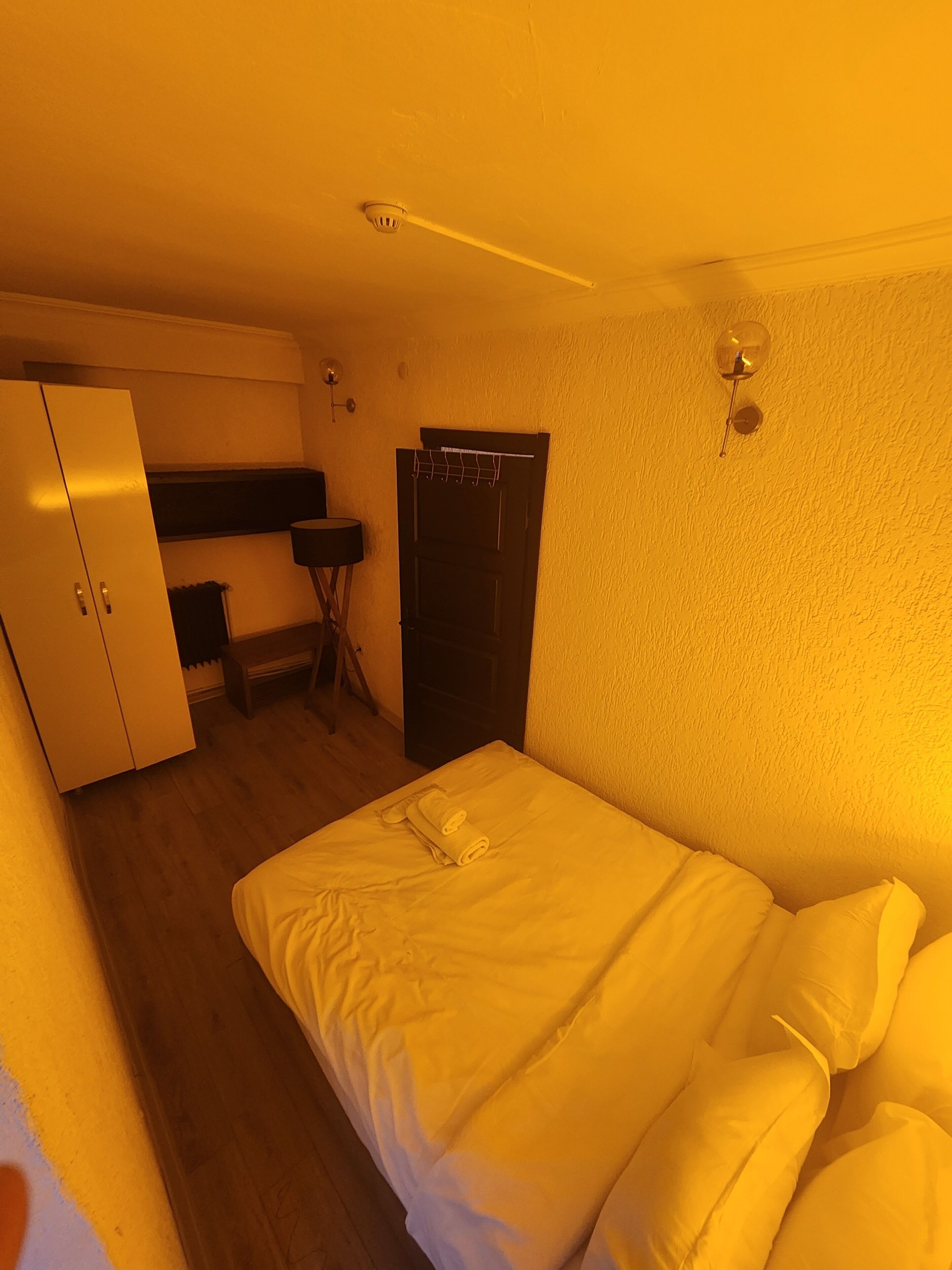 A compact hotel room with a bed, a wardrobe, a small desk, and a wall-mounted light fixture.
