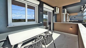 Uma varanda com mesa e cadeiras, uma janela grande com vista para o céu e nuvens, e aparelhos de ar condicionado.