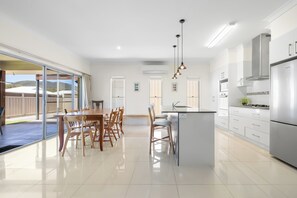 A modern kitchen with a central island, stainless steel appliances, and a dining area with wooden chairs and a glass door leading to an outdoor patio. - Sea La Vie: 4  BR, 2.5  BA House in South West Rocks, Sleeps 8 (South West Rocks)