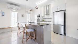 Une cuisine moderne dotée d’un îlot central, d’appareils électroménagers en acier inoxydable, de luminaires suspendus et d’une palette de couleurs épurée, dans des tons blancs.