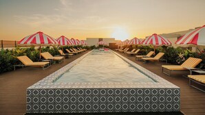 Un espace aménagé au bord de la piscine, avec des chaises longues et des parasols à rayures, une bordure de piscine décorée, et un coucher de soleil en arrière-plan.