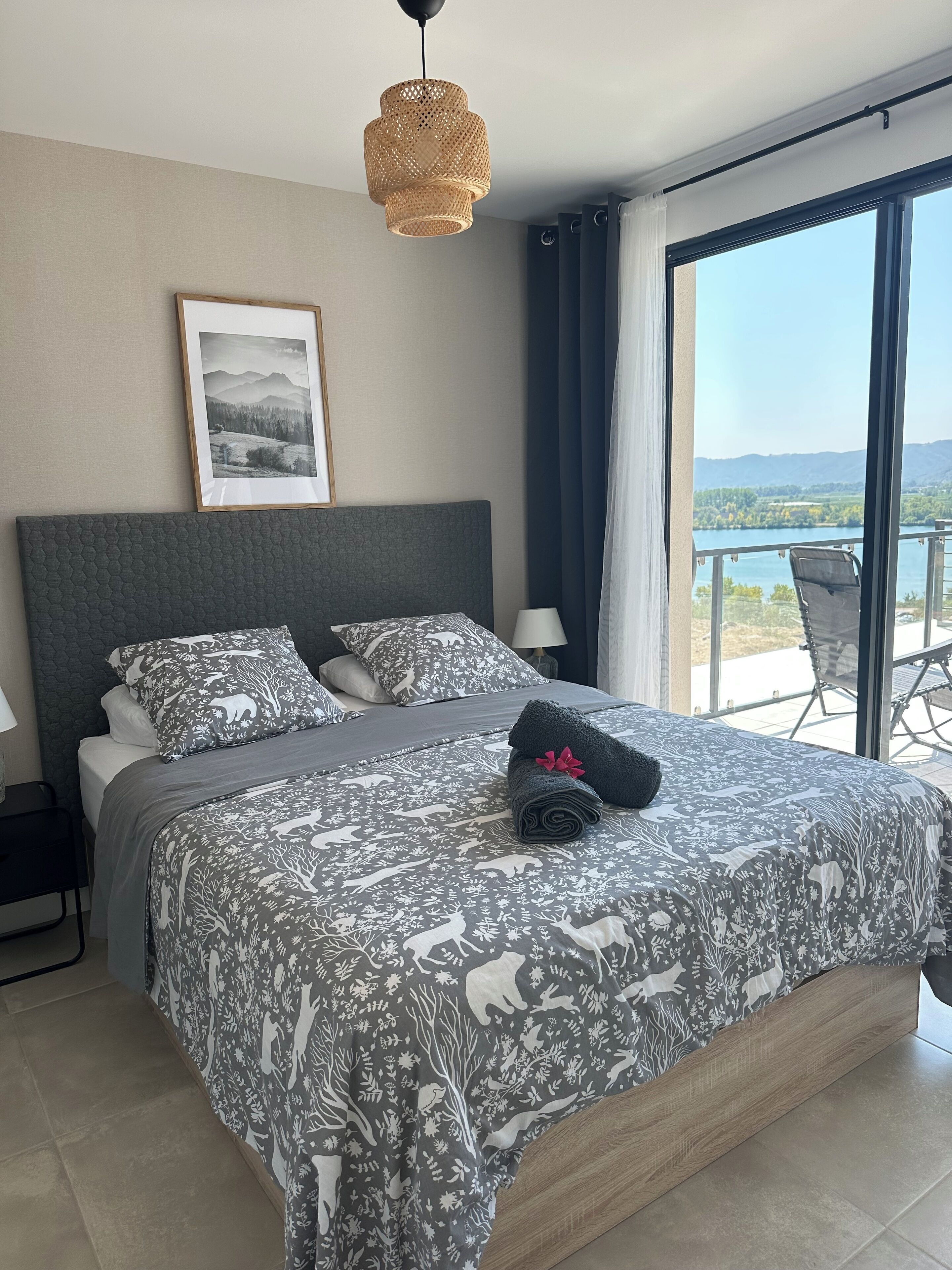 Une chambre avec un grand lit, un tableau encadré au mur et une vue sur un lac depuis un balcon.