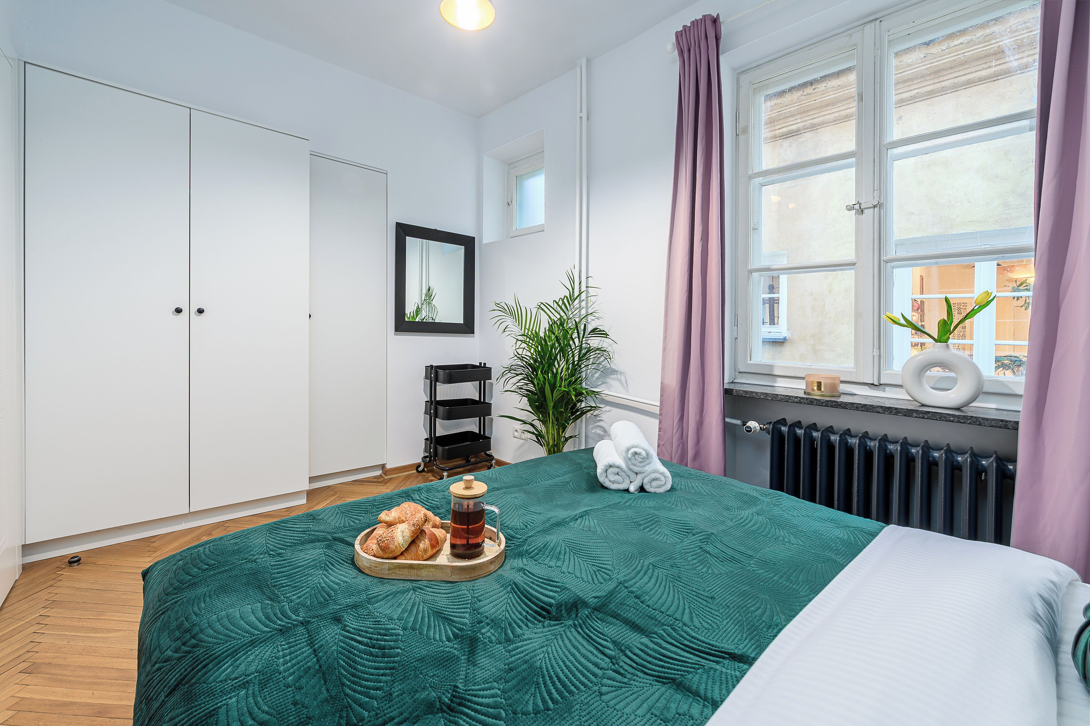 Ein ordentlich eingerichtetes Schlafzimmer mit Bett, einem Nachttisch mit Brot und Getränken, einer Topfpflanze und einem Fenster mit Vorhängen.