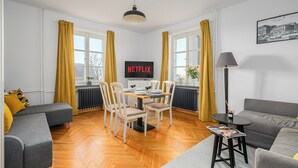 Un salon moderne comprenant un coin repas, un canapé gris, un divan gris, une table avec des chaises, un téléviseur affichant le logo « NETFLIX » et une lampe.