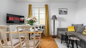 Un salon moderne avec un téléviseur affichant le logo « NETFLIX », un coin repas avec des chaises et une table, un canapé avec des coussins et une lampe sur pied.