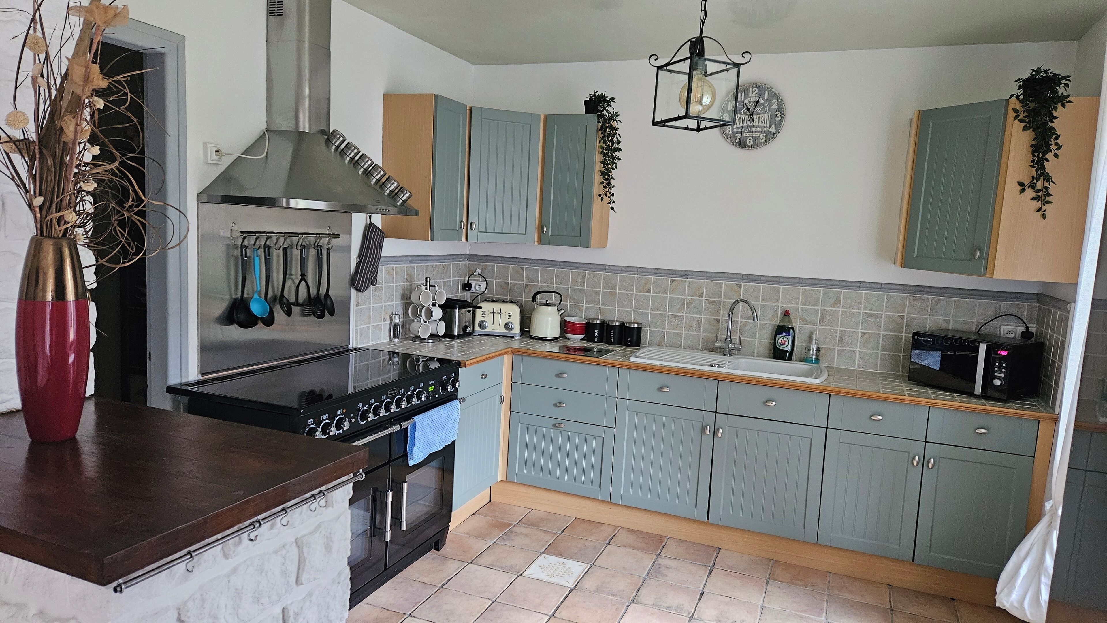 A modern kitchen with a central island, a stove, a sink, and a microwave. There are cabinets, a clock, and a hanging plant.