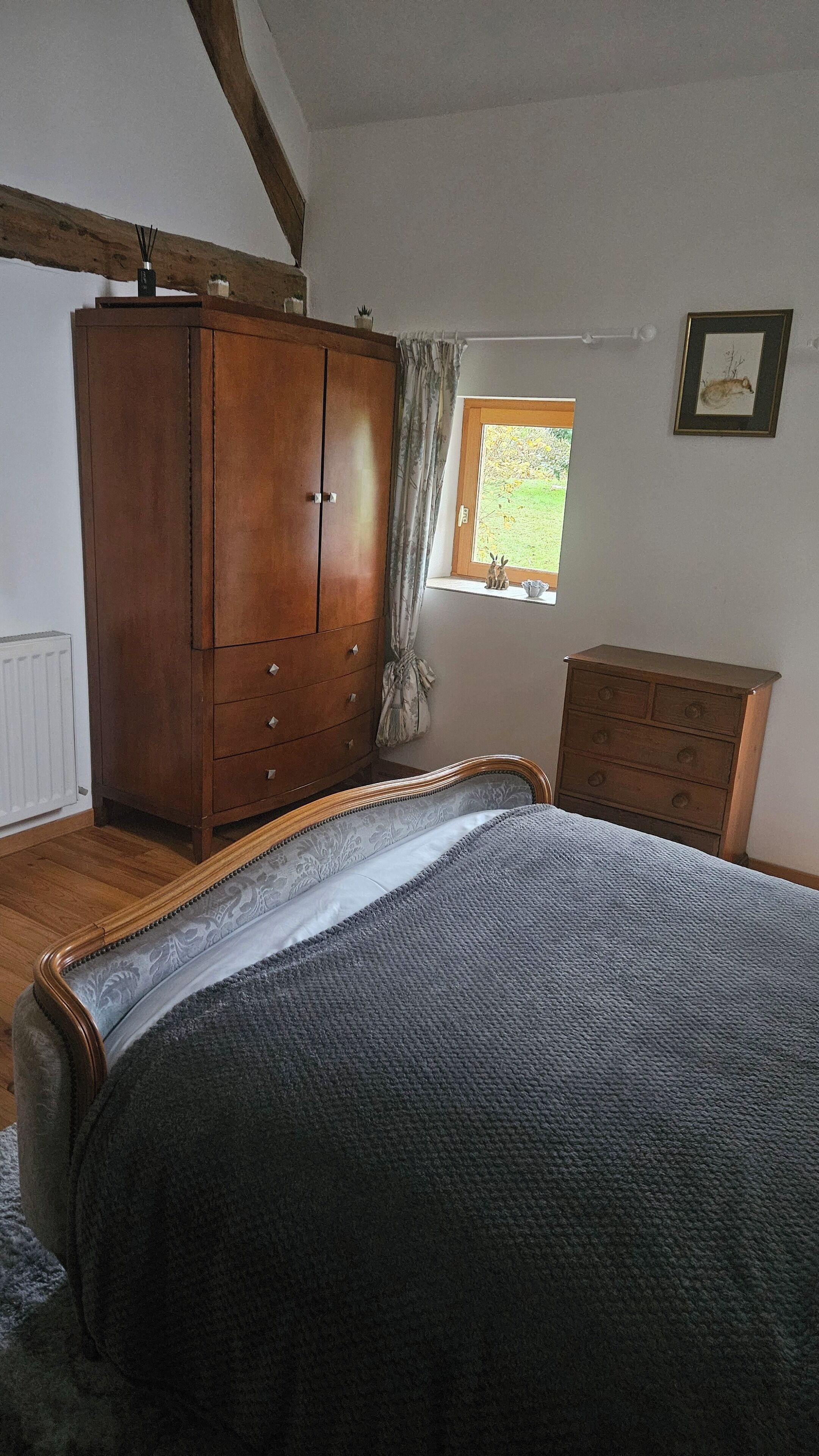 A bedroom with a wooden wardrobe, a bed with a grey blanket, a wooden dresser, and a window with a view of greenery.