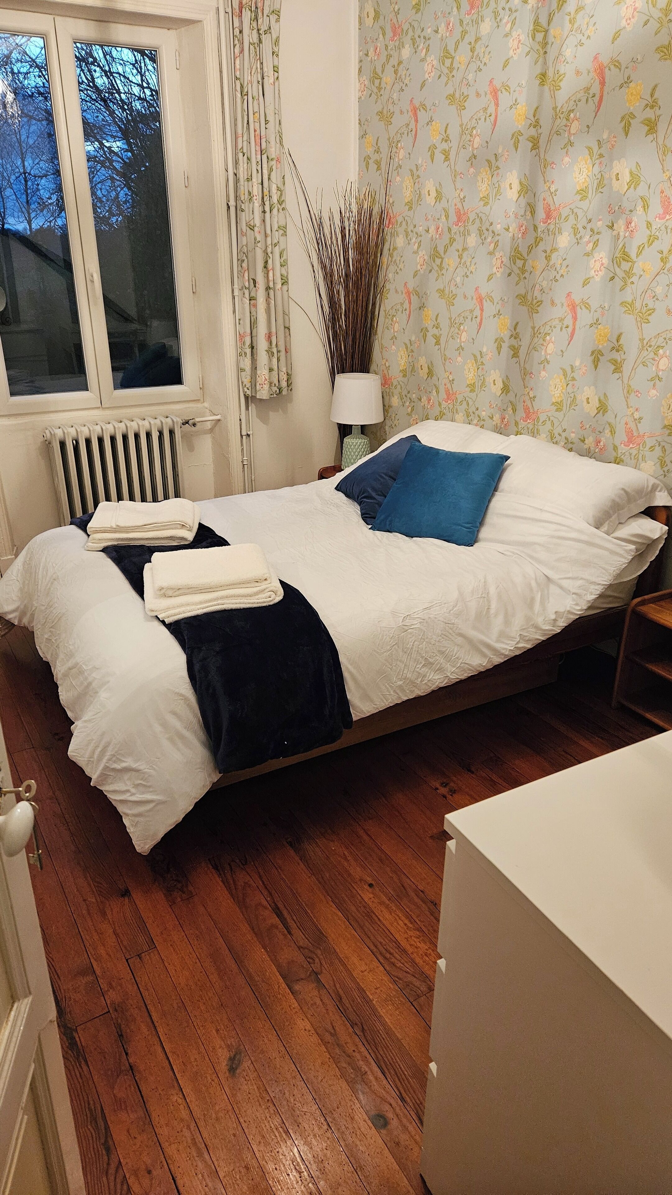 A bedroom with a wooden floor, a radiator, a window with curtains, a bed with white and blue bedding, and a floral wallpaper.