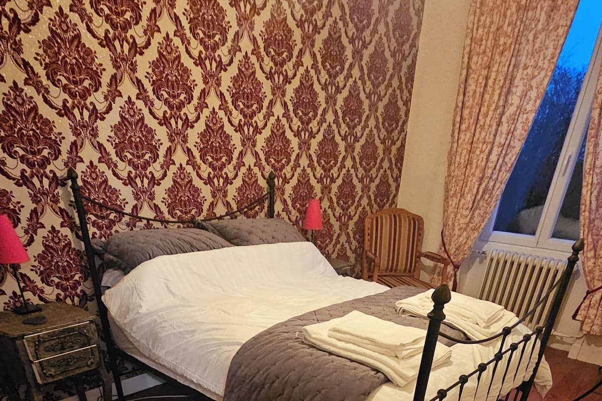 A bedroom with a wooden floor, a bed with a headboard, a nightstand, a lamp, and a window with curtains.