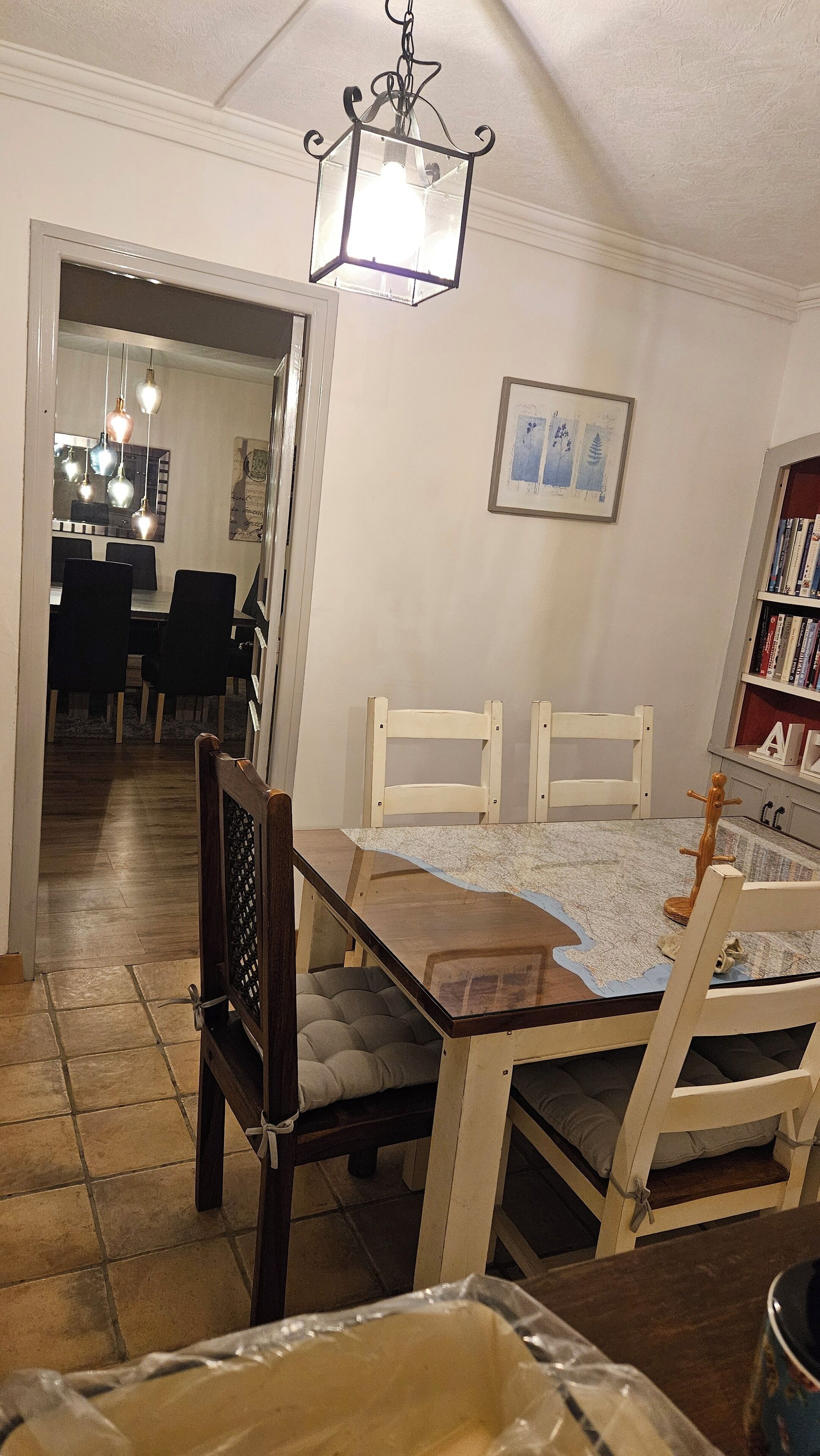 Un comedor con una mesa de madera, sillas y una araña. Hay una estantería con libros y un cuadro en la pared.