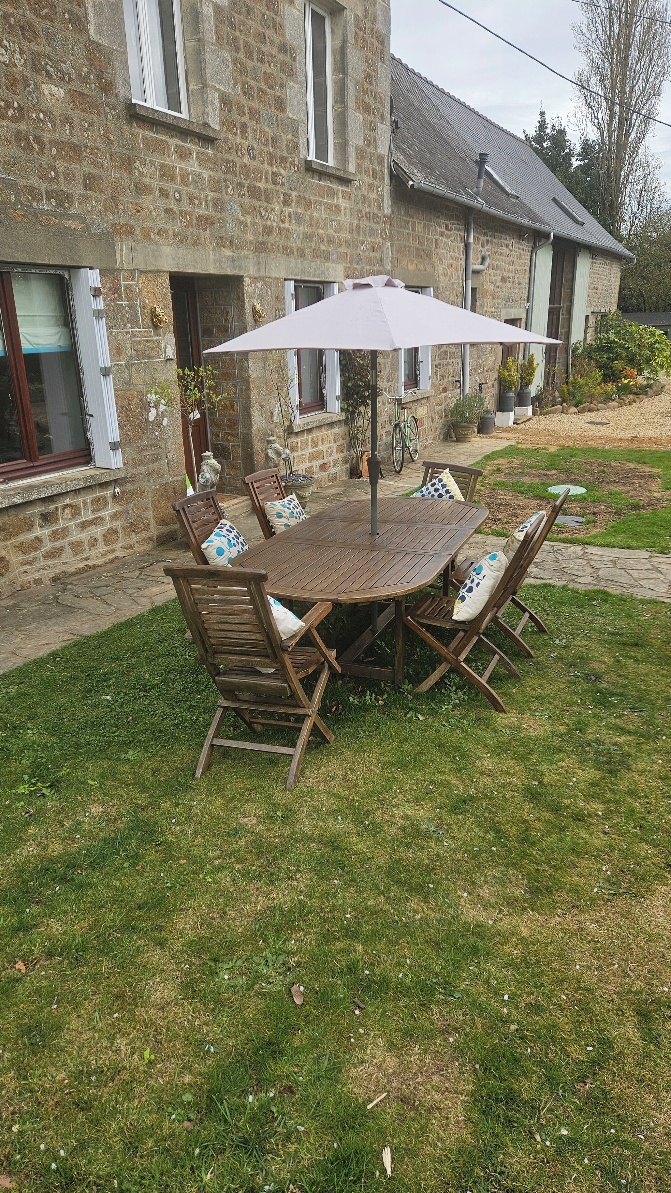 A stone house with a garden patio featuring a wooden table and chairs, an umbrella, and potted plants.