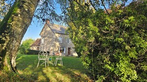 A stone house with a sloped roof, a grassy yard, and a metal table and chairs in the foreground. - The Lions Den - La Loge Mantilly (Mantilly)