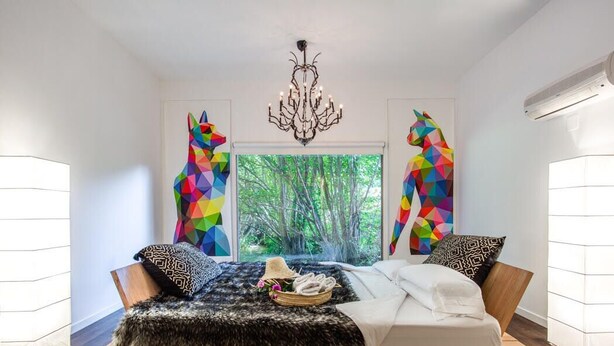 Une chambre moderne avec un lit surélevé en bois, un lustre et des œuvres d’art murales aux motifs géométriques et aux couleurs vives.