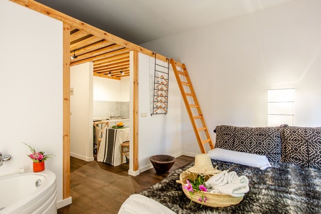 A bedroom with a wooden ceiling, a bed with patterned pillows, a small bathroom with a sink and a plant, and a ladder against the wall.