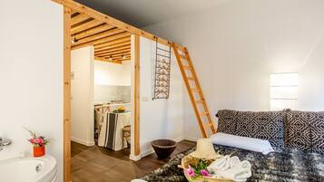 A bedroom with a wooden ceiling, a bed with patterned pillows, a small bathroom with a sink and a plant, and a ladder against the wall.