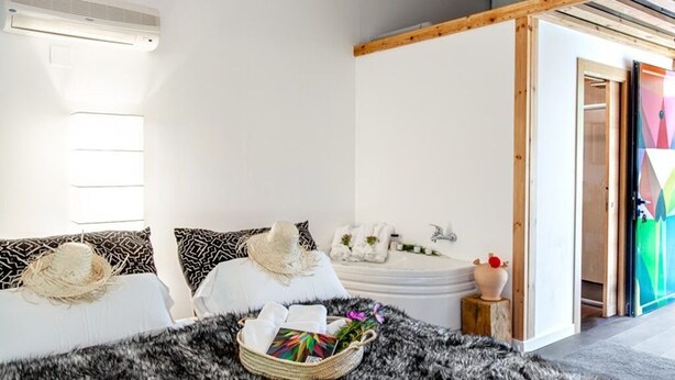 Une chambre moderne avec un grand lit, une petite table de chevet, un panier contenant des articles de toilette et une vue sur une salle de bain avec une baignoire.