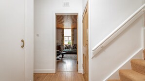 A hallway with a wooden floor, a staircase with carpeted steps, and a view into a living room with a sofa and a window.