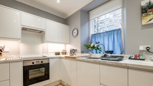 A modern kitchen with white cabinets, a built-in oven, a window with blue curtains, and a clock on the wall.