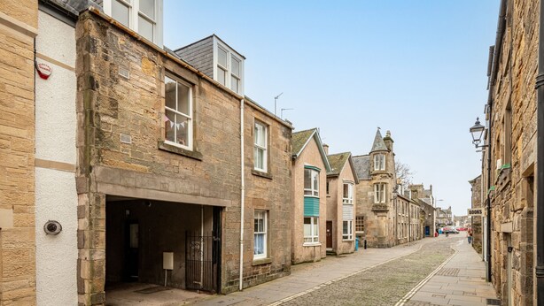 A narrow cobblestone street lined with historic stone buildings, some with visible shop fronts and signage.