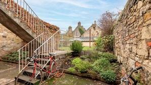 A narrow alley with a red bicycle leaning against a white railing, stone walls, and a small garden area with plants and a pathway.