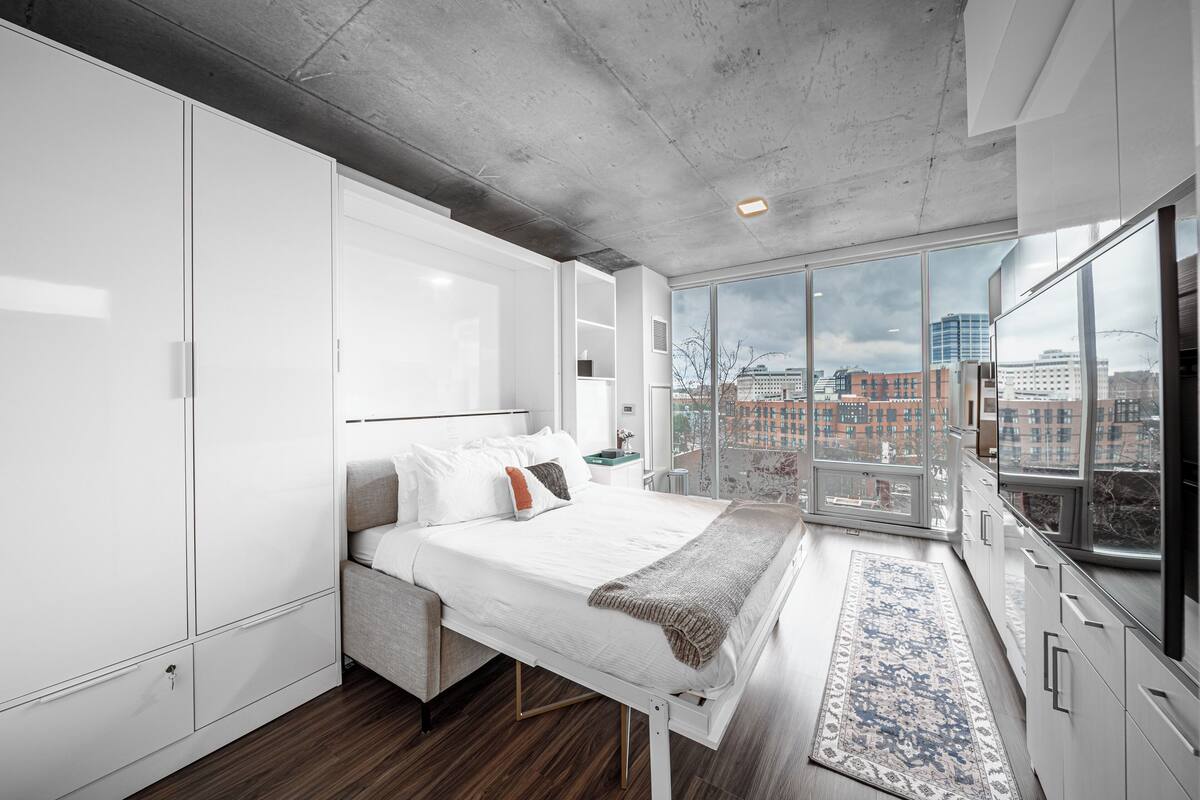 A modern bedroom with a large bed, white cabinetry, and a view of the city through floor-to-ceiling windows.