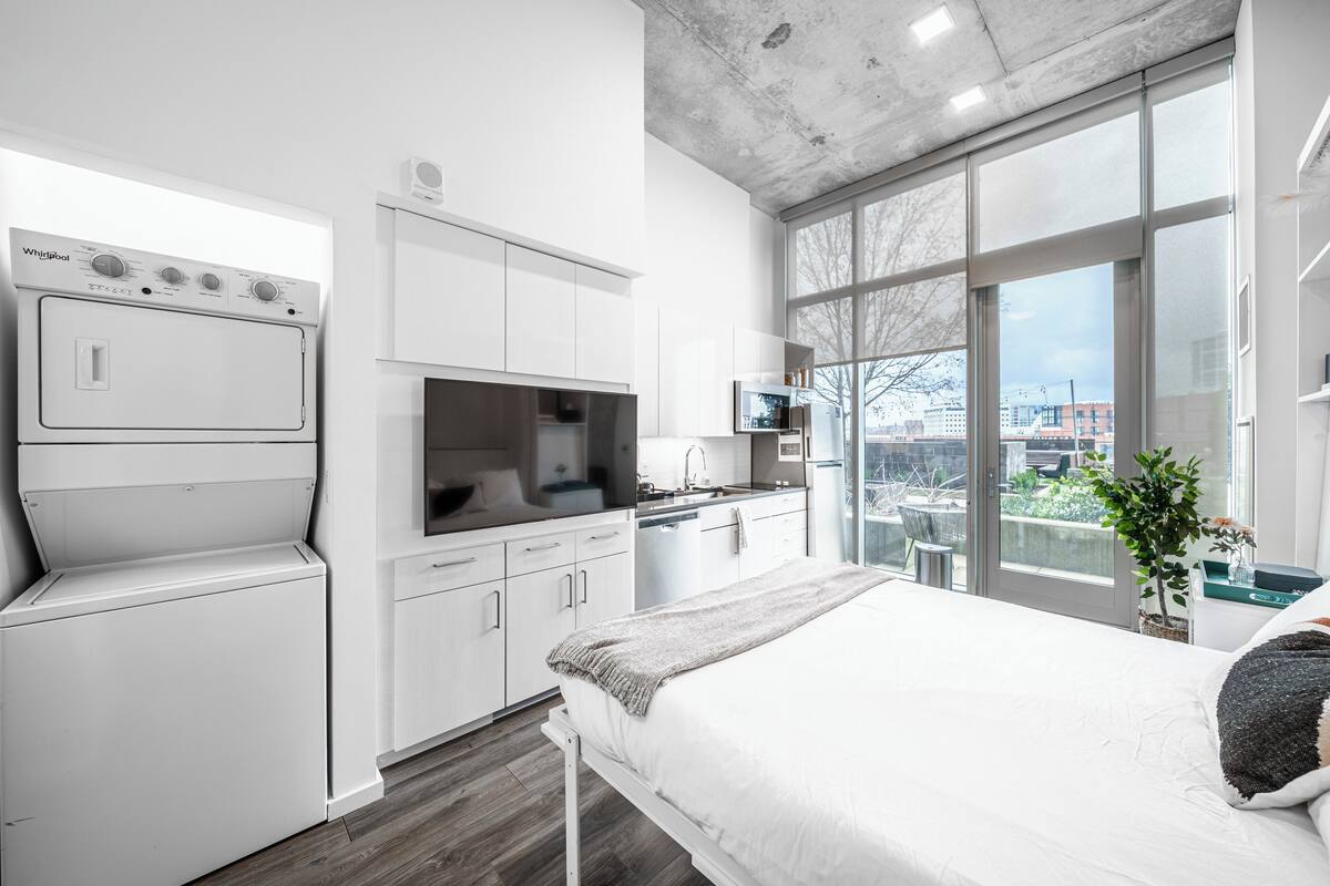 A modern bedroom with a large bed, a built-in washer and dryer, a kitchenette, and a large window offering a view of the city.