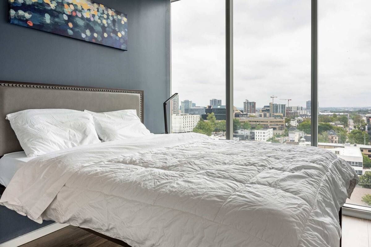 A bedroom with a large bed, a headboard, and a city view through a large window.
