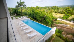Piscina moderna con sillones blancos, rodeada de exuberante vegetación y vistas a colinas distantes.