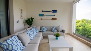 Una sala de estar moderna con un sofá beige, cojines estampados, una mesa de centro blanca y vistas a un follaje exuberante a través de amplios ventanales.
