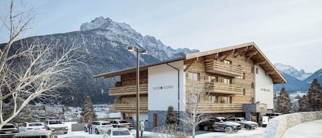A modern hotel with a wooden facade, named 'Suites Fulpmes', featuring a multi-story structure and a parking area with several cars.