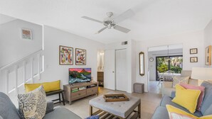 Ein Wohnzimmer mit grauem Sofa, einem Couchtisch und einem Fernseher. An den Wänden befinden sich Gemälde und es gibt einen Deckenventilator.