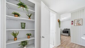 A hallway with white shelves holding potted plants, a white door, and a kitchen area with a stove and a fire extinguisher. - Stay just minutes from Downtown in this cozy, well-designed 1-bedroom apartment. An ideal base for professionals, students, and travelers wanting fast access to Houston’s best dining and attractions. (Houston)