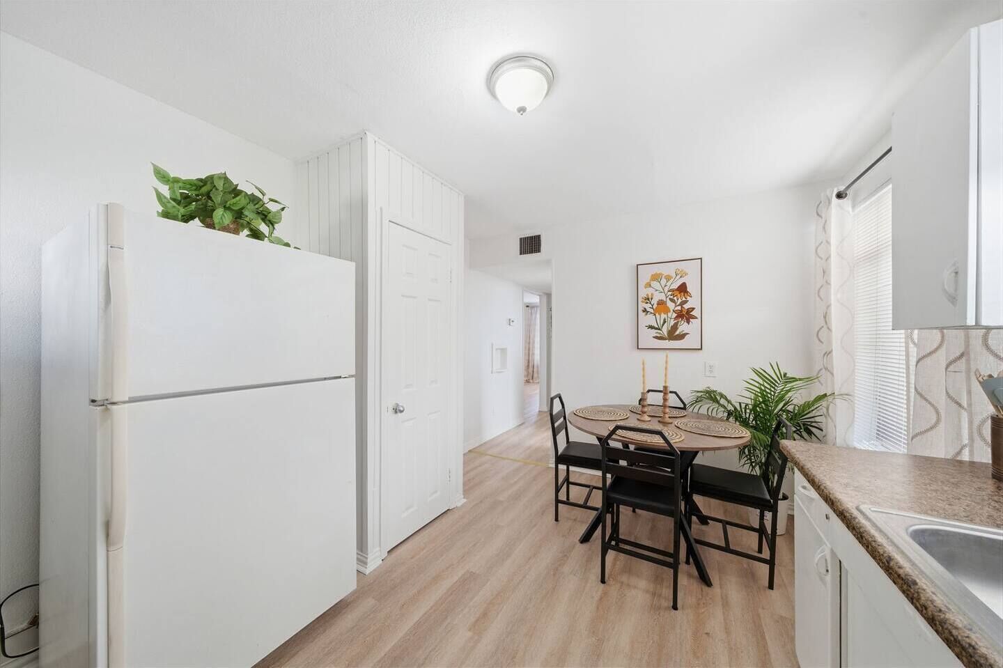 A compact kitchen with a white refrigerator, a dining table with chairs, and a small window with curtains.