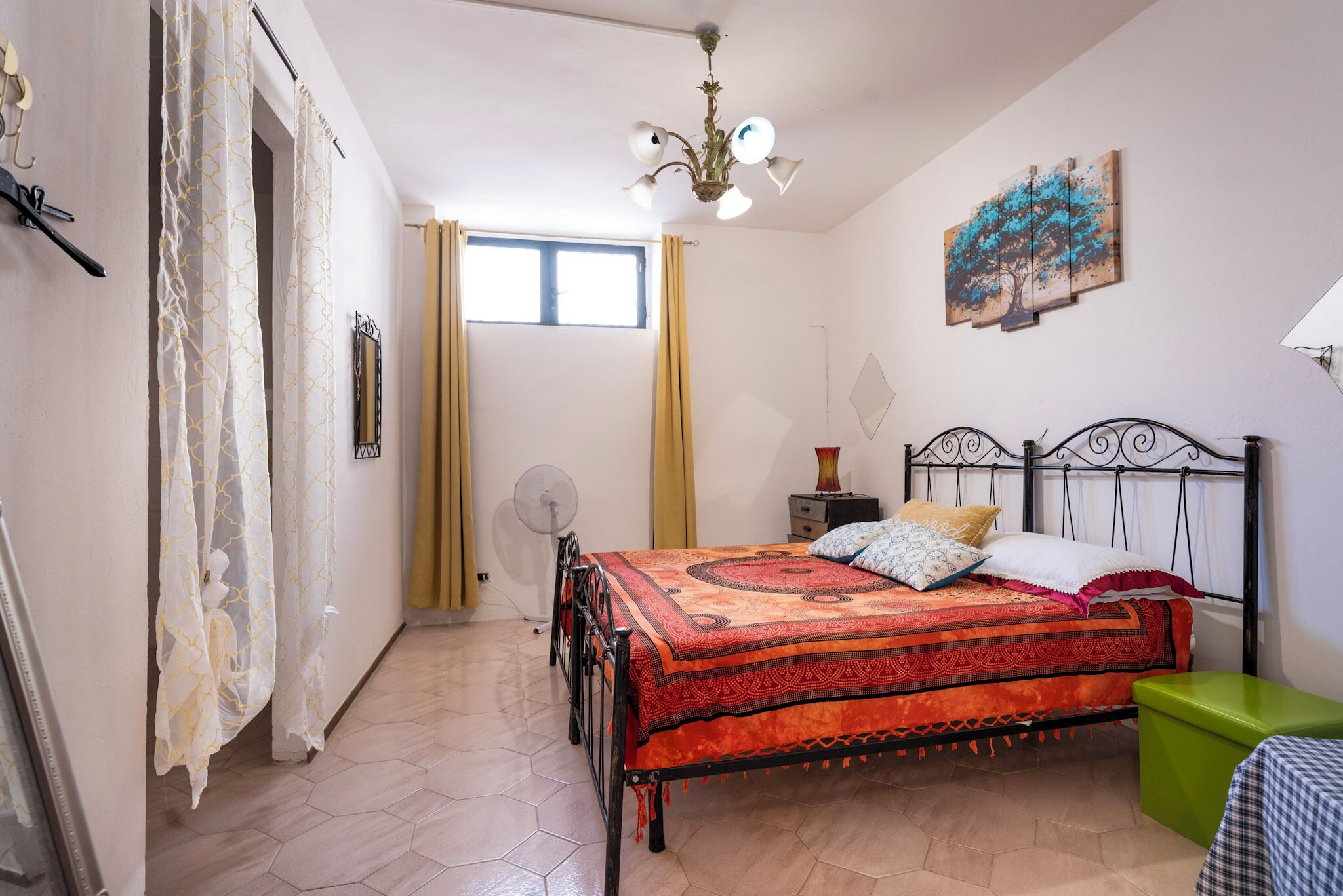 A bedroom with a bed, a fan, a green stool, and a painting on the wall.