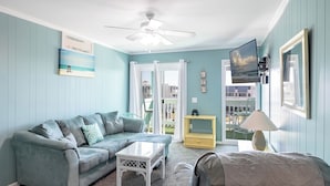 A living room with a light blue wall, a grey sofa, a white coffee table, and a ceiling fan. - SeaSpray 312, Oceanview Condo in Atlantic Beach, NC! (Atlantic Beach)