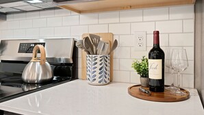 En köksbänk med en vattenkokare i rostfritt stål, ett förvaringsställ för köksredskap, en vinflaska och glas.