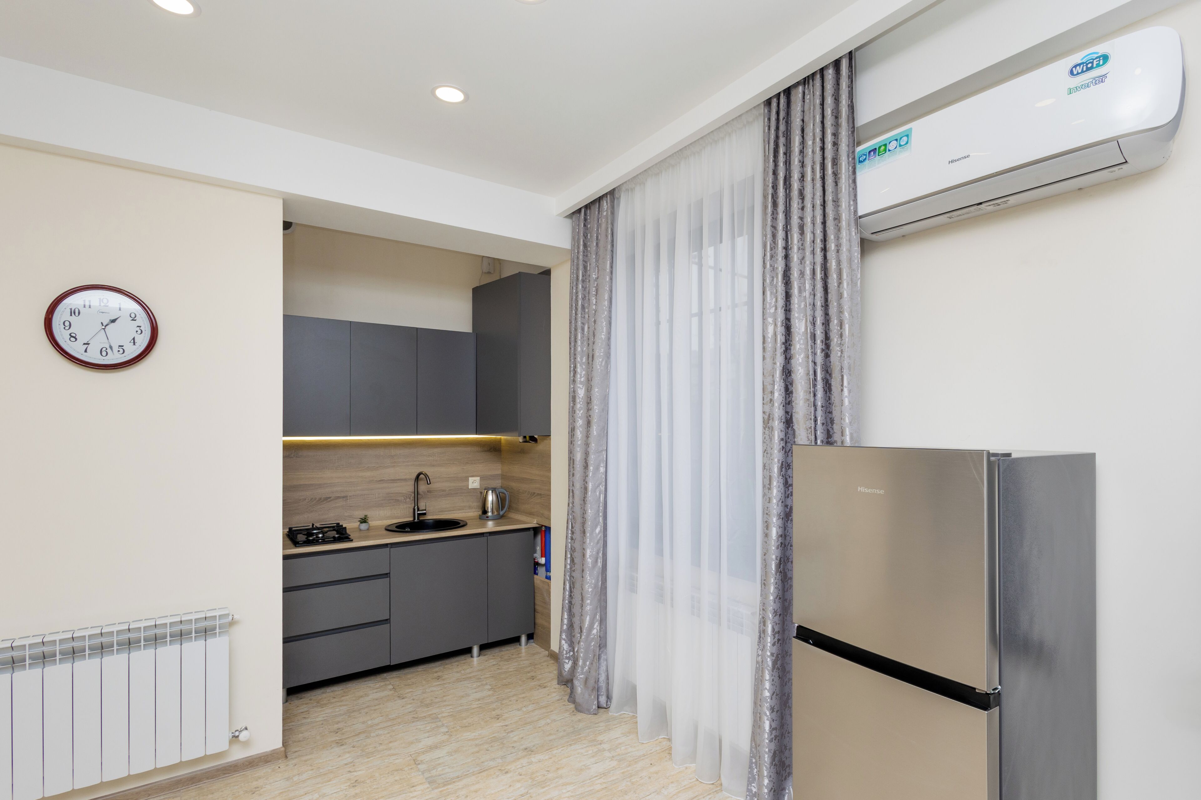 A modern kitchen with grey cabinets, a stainless steel refrigerator, a clock, and an air conditioning unit.