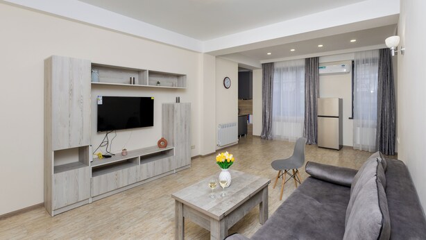 A modern living room with a flat-screen TV, grey sofa, and a coffee table with a vase of flowers. - Apartment by Luxe Group XO222 (Yerevan)