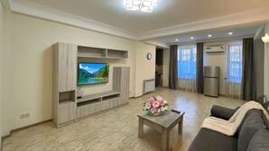 A modern living room with a flat-screen TV, wooden entertainment unit, a coffee table with flowers, a sofa, and a kitchen area with a refrigerator and oven. - Apartment by Luxe Group XO222 (Yerevan)