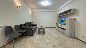 A modern living room with a grey sofa, two armchairs, a coffee table with flowers, a flat-screen TV on a wooden TV stand, and recessed ceiling lighting. - Apartment by Luxe Group XO222 (Yerevan)
